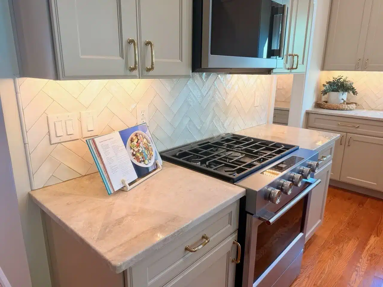 Stainless steel gas range under a microwave, flanked by gray shaker cabinets with gold pulls, with a glossy white herringbone tile backsplash and under-cabinet lighting