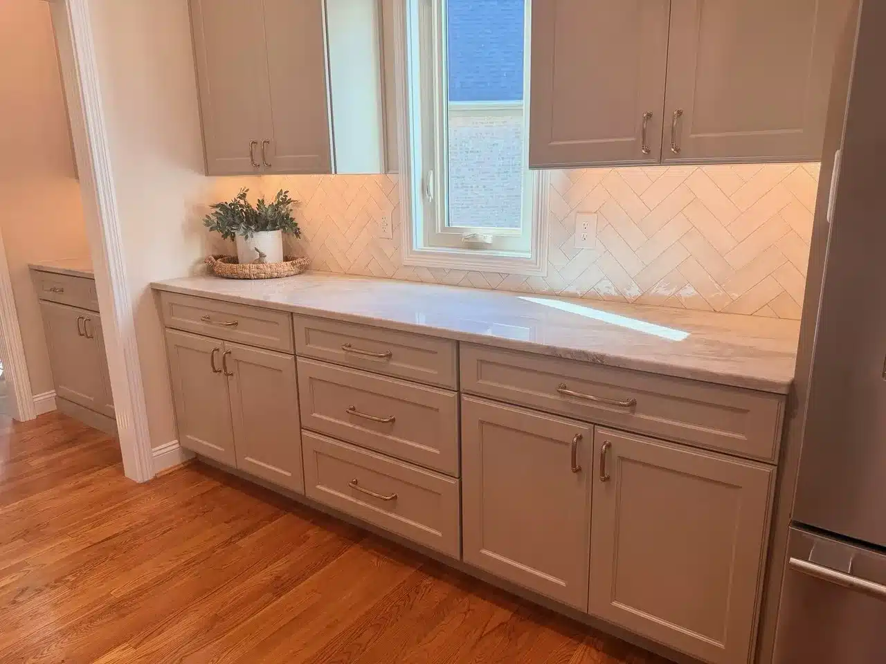 Gray shaker base and wall cabinets with brushed gold pulls, a white marble-look countertop and a white herringbone tile backsplash beside a window